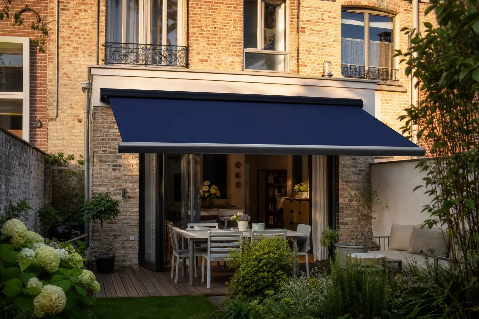 Maison de ville en brique typique de la métropole lilloise avec un store banne à coffre bleu marine déployé sur la terrasse, jardin urbain, lumière de fin de journée